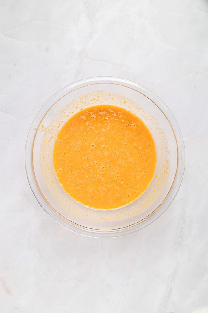A glass bowl filled with a whisked yellow-orange mixture for Sweet Potato Cornbread sits on a white marble surface.