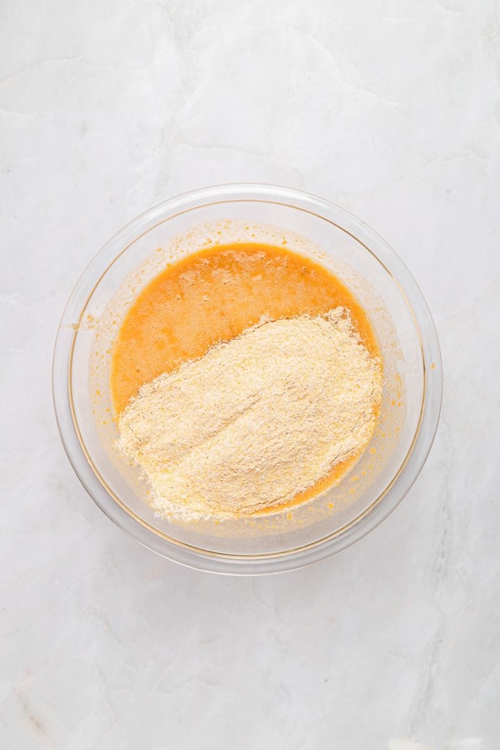 A glass bowl with Sweet Potato Cornbread batter and dry cornmeal sits on a white marble surface.