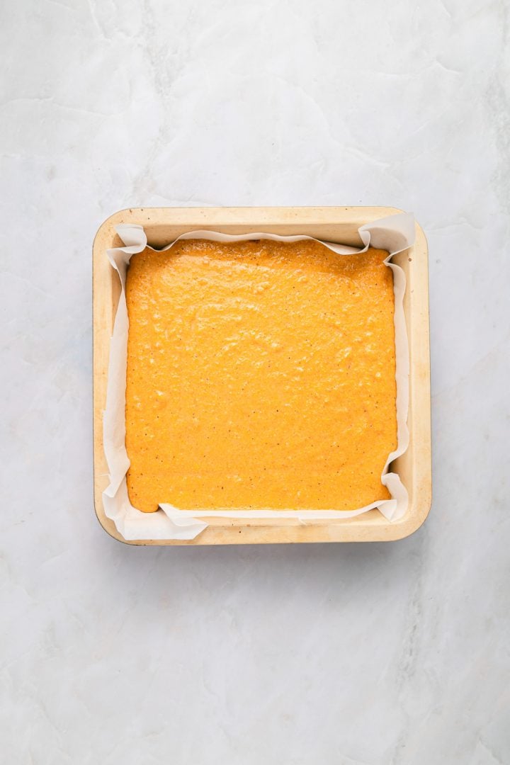 Square baking pan lined with parchment paper filled with sweet potato cornbread batter, on a light marble surface.