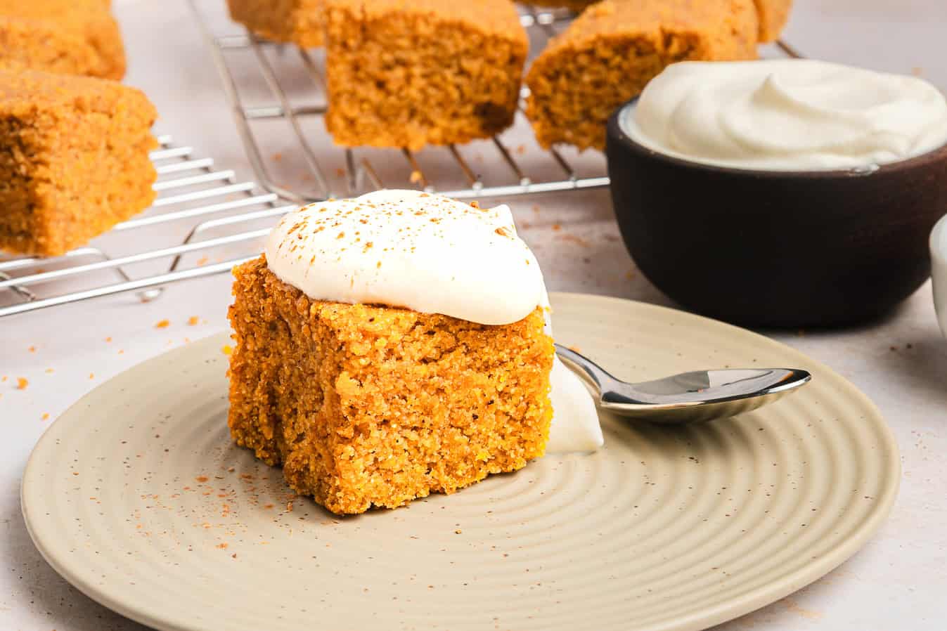 A slice of sweet potato cornbread topped with whipped cream on a plate, with more pieces and cream in the background.