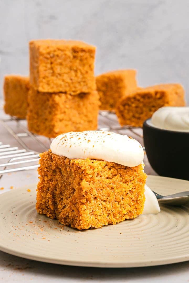 A slice of spiced carrot cake with cream cheese frosting sits on a plate, with more cake squares and sweet potato cornbread in the background.