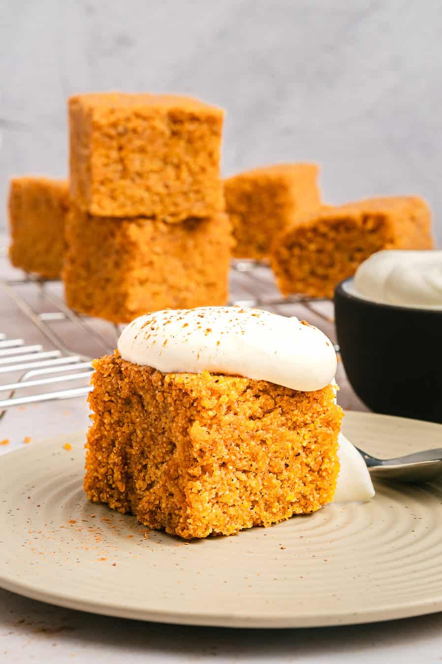 A slice of spiced carrot cake with cream cheese frosting sits on a plate, with more cake squares and sweet potato cornbread in the background.