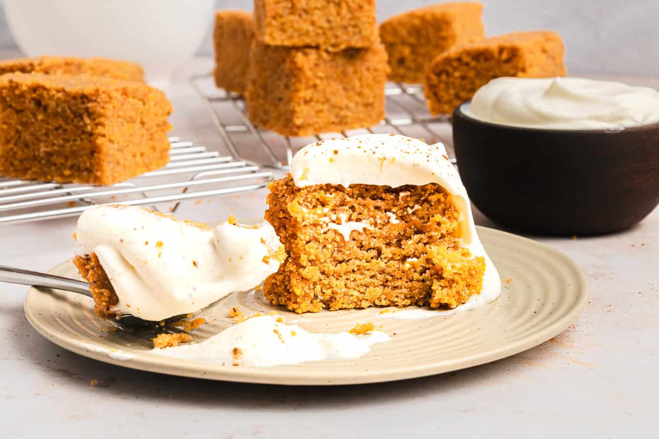 A slice of spiced cake with frosting sits on a plate beside a fork, with extra Sweet Potato Cornbread squares in the background.