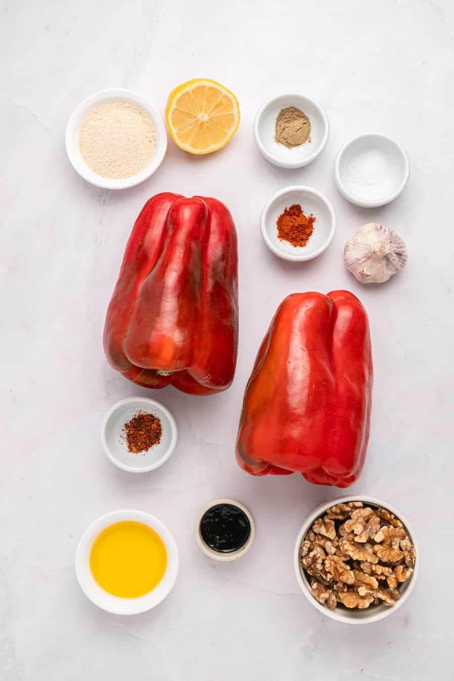 Two red bell peppers with small bowls of spices, oil, walnuts, lemon, and garlic on a white surface.