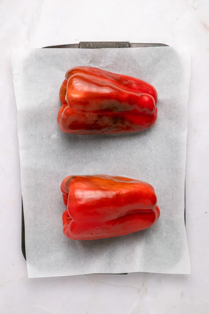 Two red bell peppers on a parchment-lined baking sheet, ready for roasting.