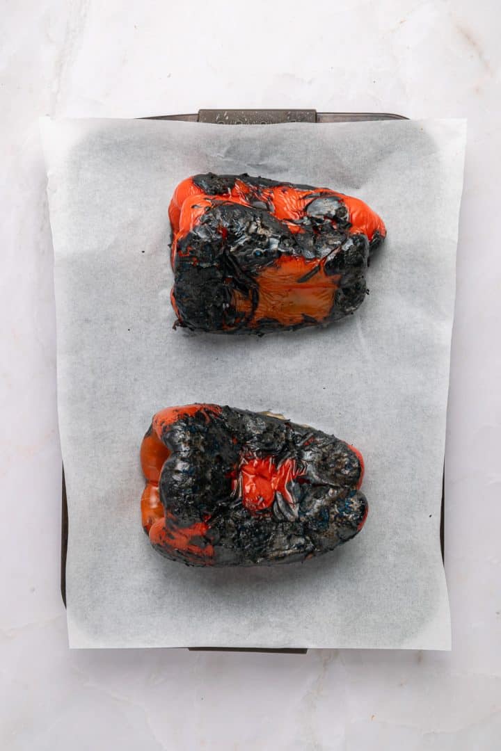 Two charred red bell peppers on parchment paper atop a baking tray, photographed from above.
