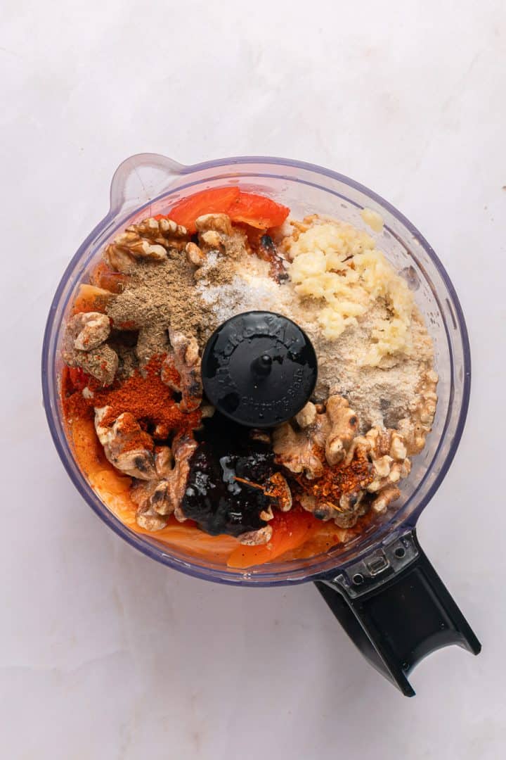 Food processor with walnuts, spices, chopped tomatoes, and minced garlic on a white surface, ready to blend.