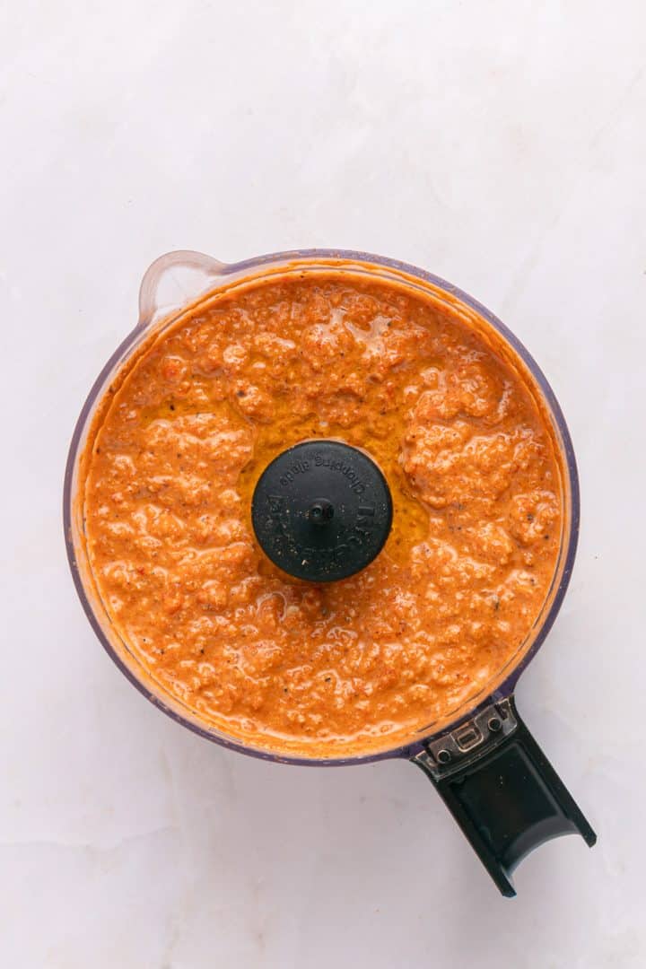 Orange-red sauce with a textured consistency in a food processor, viewed from above on a white surface.