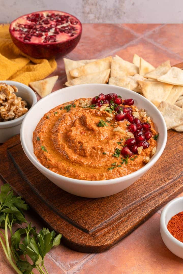A bowl of red pepper dip topped with pomegranate seeds and walnuts, served with pita chips and herbs.