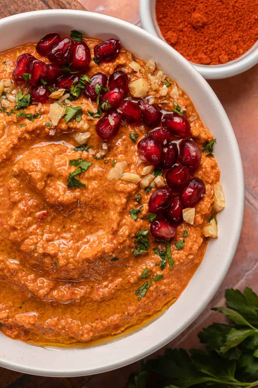Red pepper dip garnished with pomegranate seeds, chopped nuts, and herbs in a white bowl.