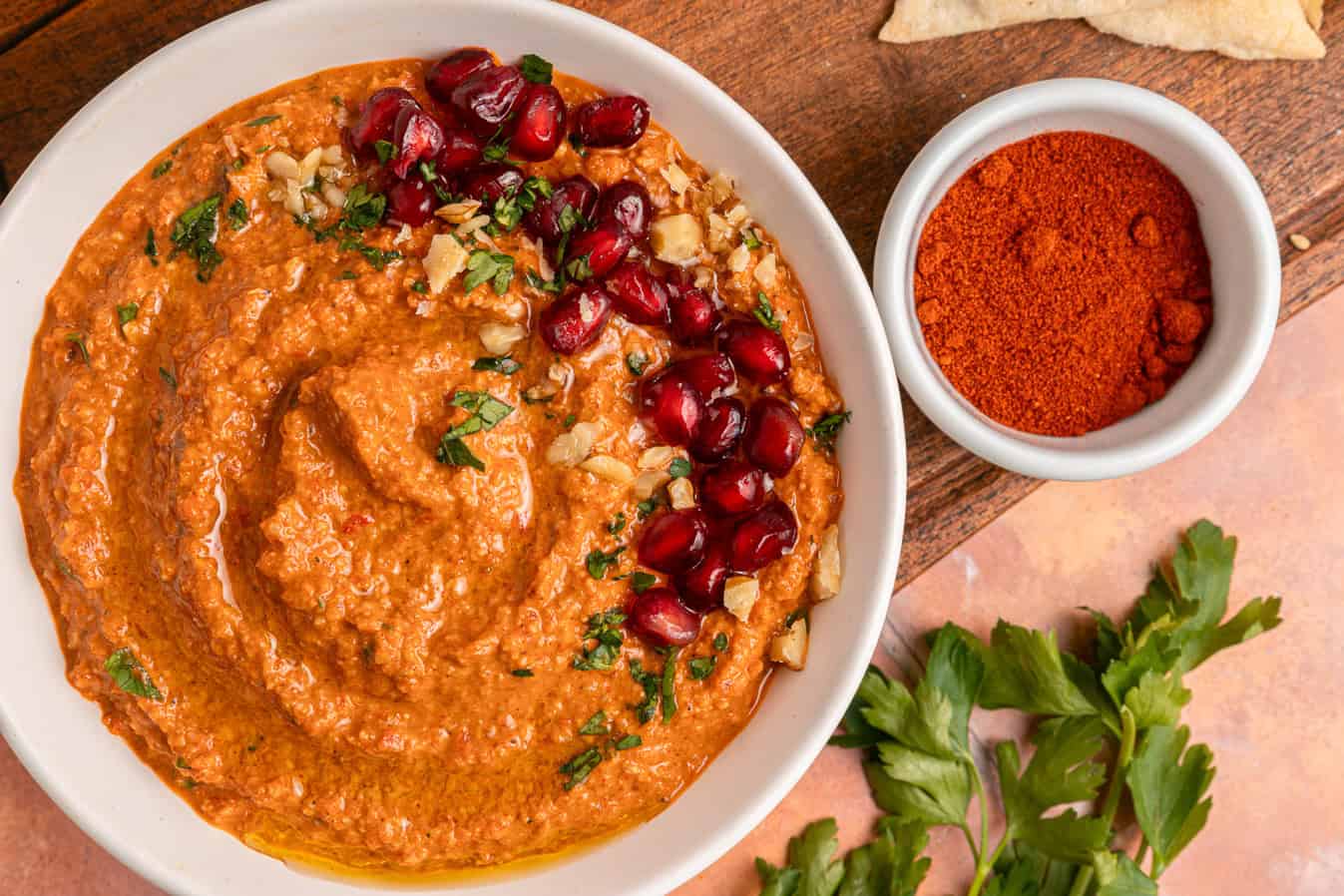 Bowl of muhammara topped with pomegranate seeds, herbs, and walnuts, beside paprika and parsley.