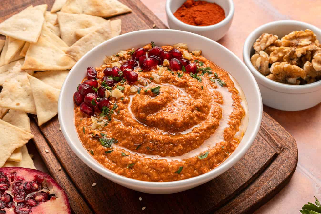 A bowl of muhammara dip topped with pomegranate seeds and walnuts, with pita chips, walnuts, and spices on the side.