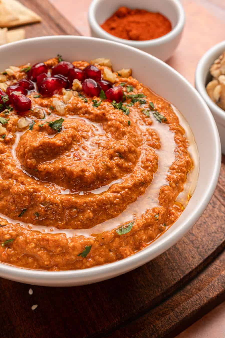 A bowl of muhammara dip garnished with pomegranate seeds, walnuts, and herbs on a wooden board.