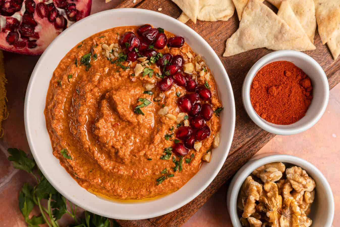 A bowl of muhammara dip topped with pomegranate seeds, surrounded by pita chips, paprika, and walnuts.