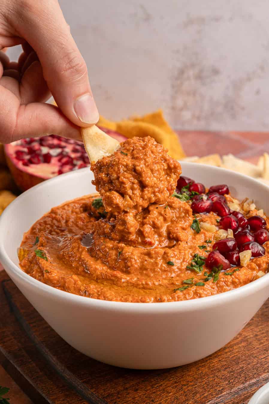 A hand dips pita bread into a bowl of red dip, garnished with herbs and pomegranate seeds.