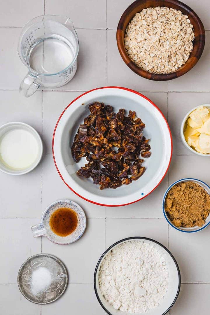 A variety of baking ingredients in bowls, including oats, dates, flour, brown sugar, butter, milk, and vanilla.