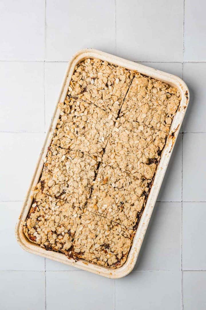 A rectangular baking dish filled with oat crumble bars, divided into pieces, on a light tiled surface.