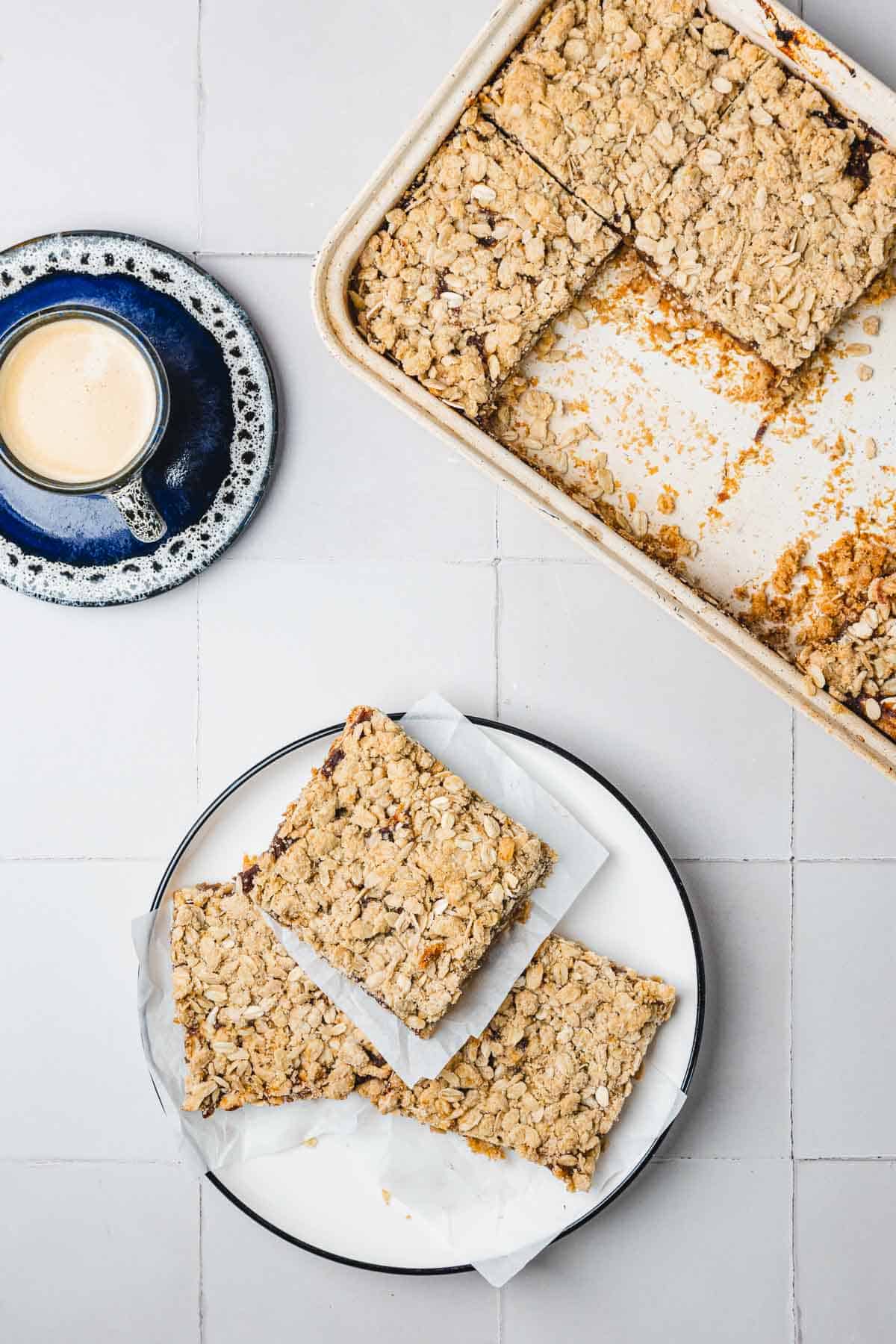 A plate with two oat bars on parchment paper, a tray of bars, and a cup of coffee on a tiled surface.