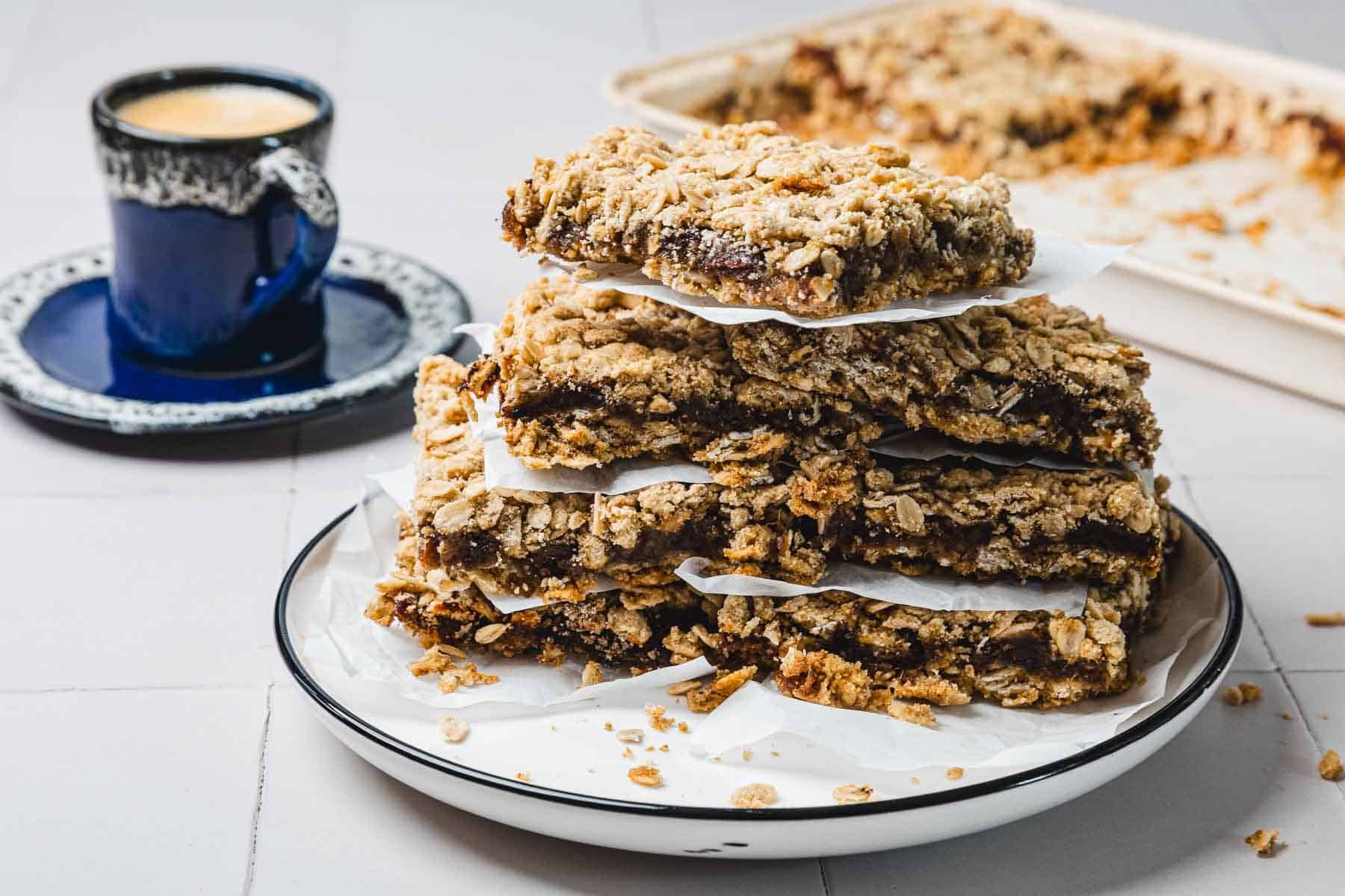A stack of oatmeal crumble bars on a plate, with a cup of coffee and a baking tray in the background.