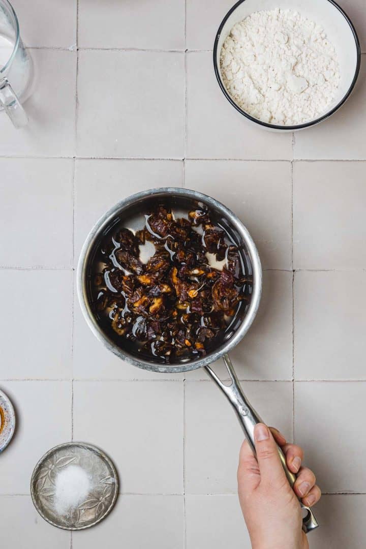 A hand holds a saucepan with chopped dates soaking in water, next to a bowl of flour and a small dish of sugar.