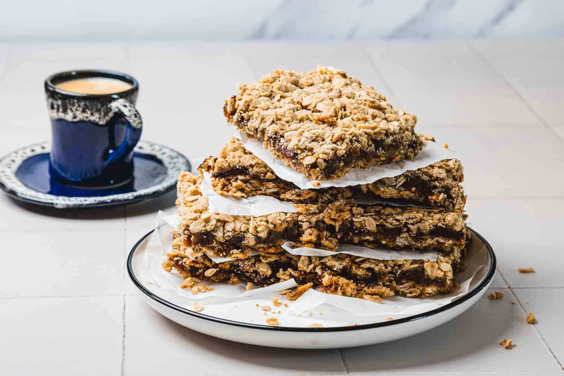 A stack of crumbly oat bars on a plate with a cup of coffee in the background.