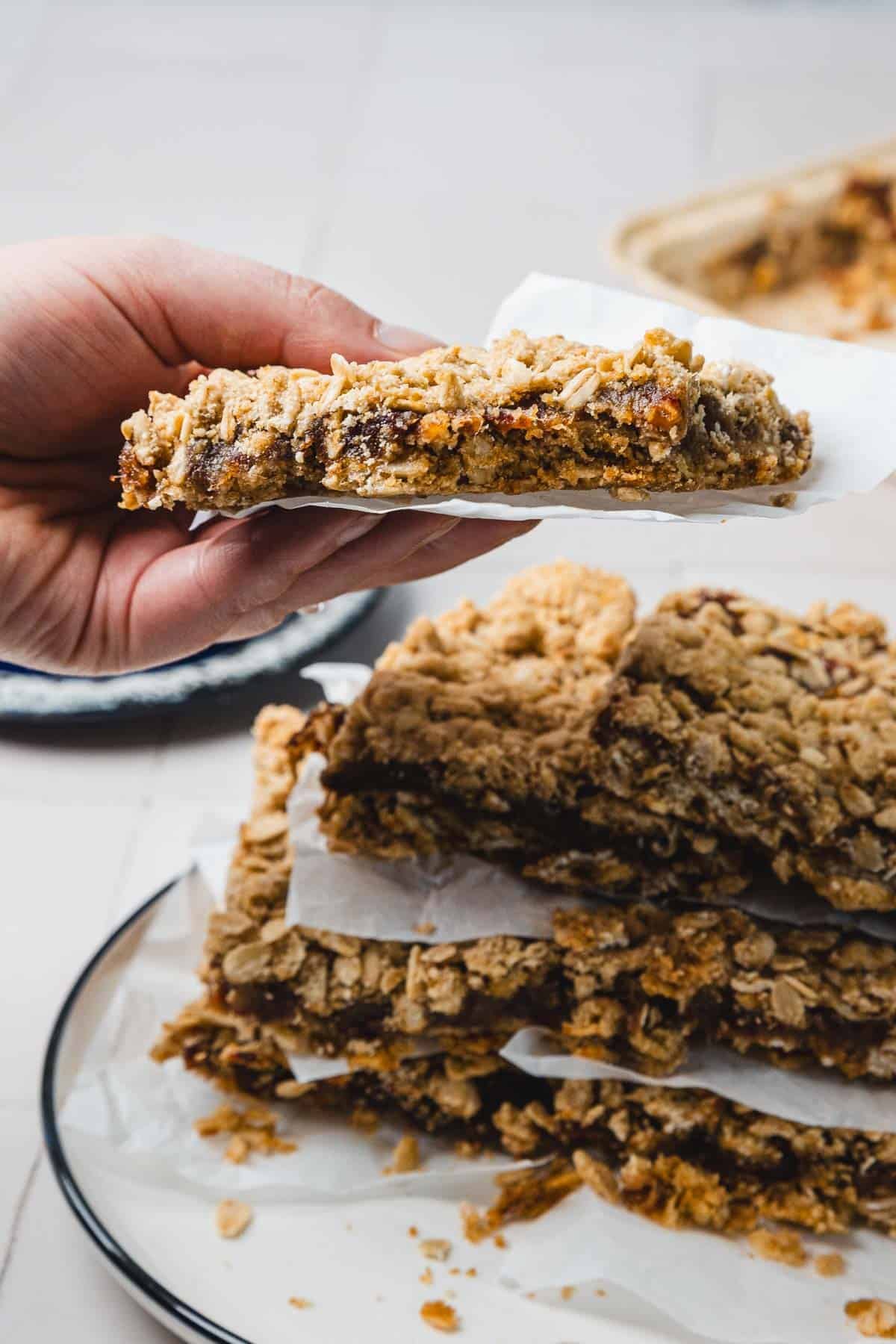 A hand holds an oat bar over a plate stacked with more oat bars, separated by parchment paper.