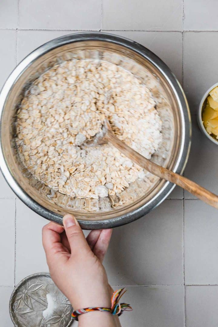 A hand mixes oats and flour in a metal bowl with a wooden spoon; butter is in a bowl nearby.
