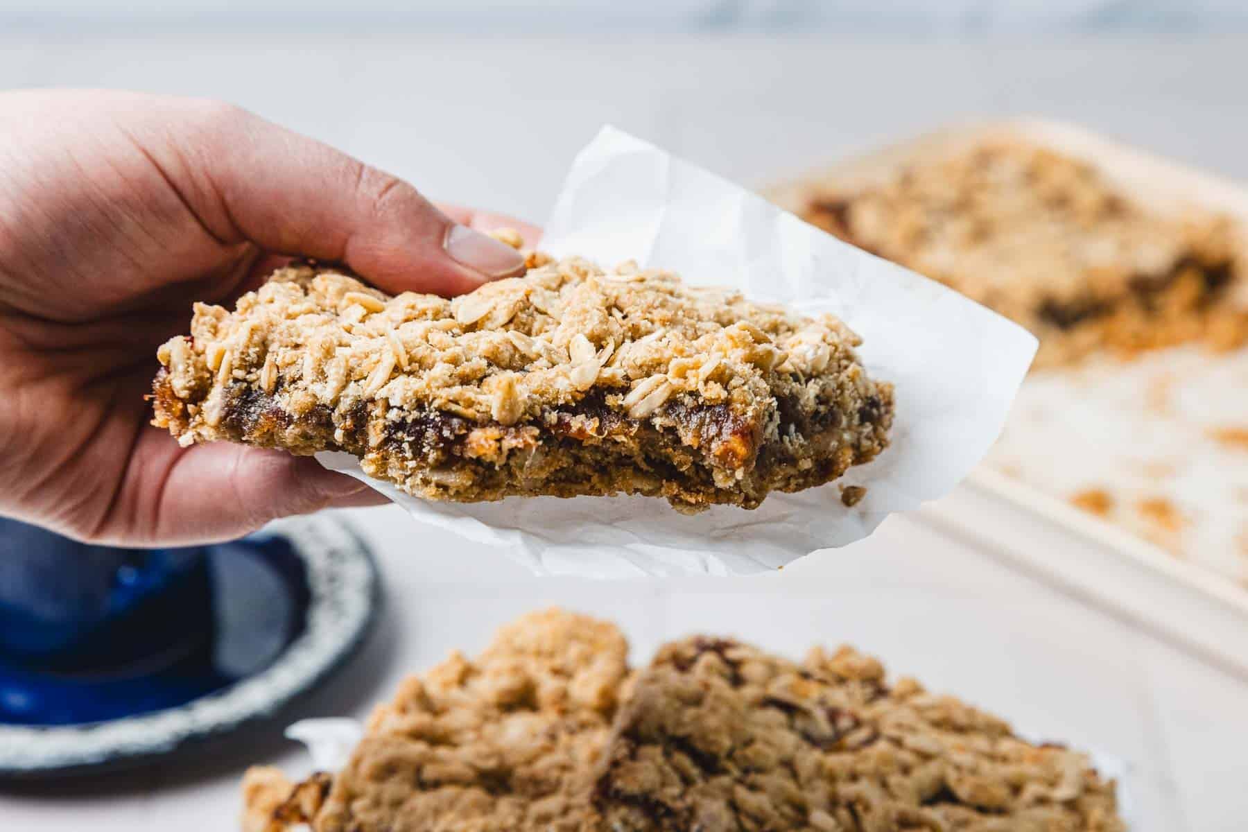 A hand holding a crumbly oat bar with a fruit filling, wrapped in white parchment paper.