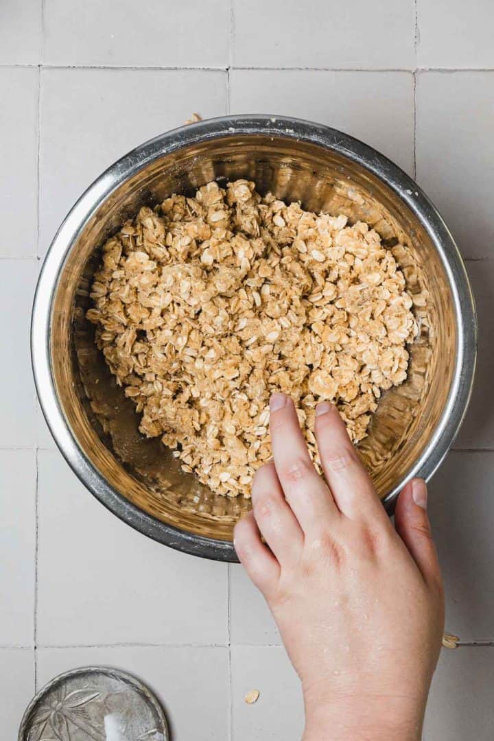 A hand mixing oats in a stainless steel bowl on a light tiled surface.