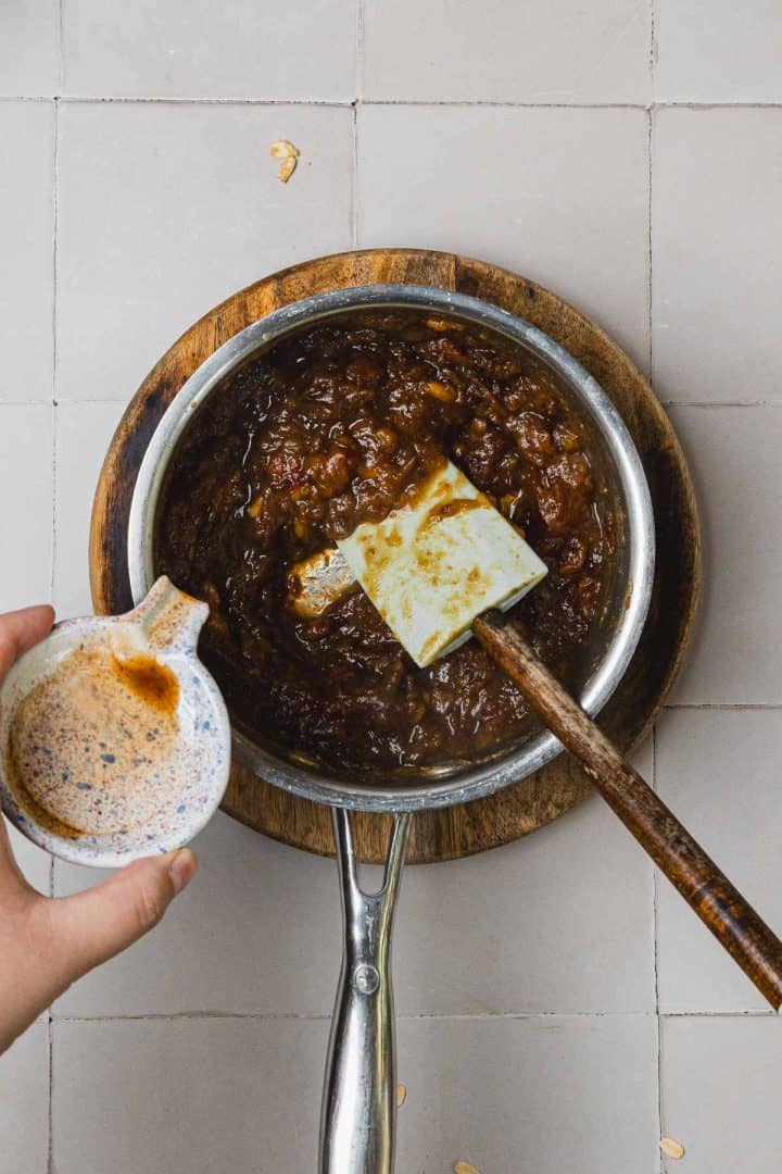 A hand pours liquid into a saucepan of dark chutney with a spatula, on a wooden cutting board.