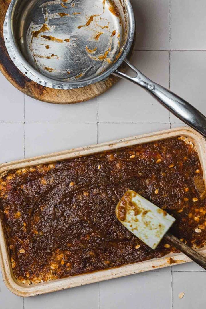 A spatula rests on a pan of date paste next to an empty, messy metal saucepan on a tiled counter.