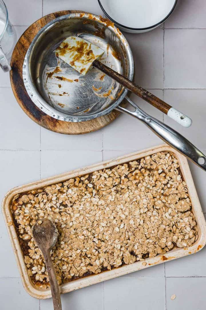 A baking dish with oat crumble dessert, a wooden spoon, and empty mixing bowls on a tiled countertop.
