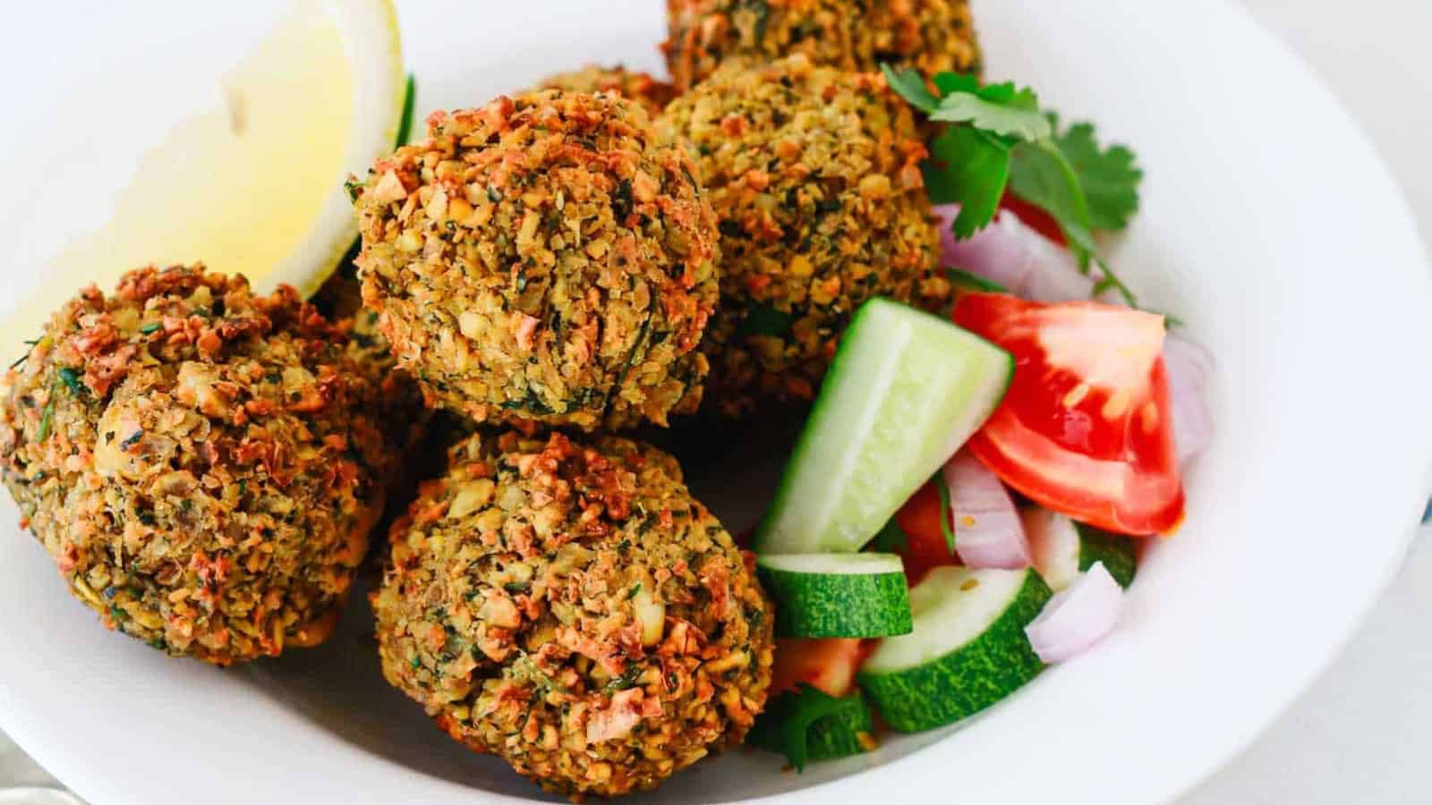 Four falafel balls with lemon wedge, cucumber, tomato, onion, and cilantro on a white plate.