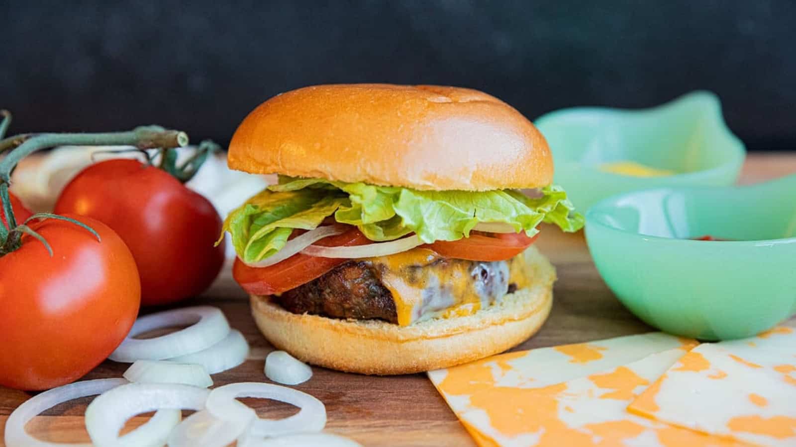 A cheeseburger with lettuce and tomato next to sliced onions, cheese, tomatoes, and green bowls on a board.