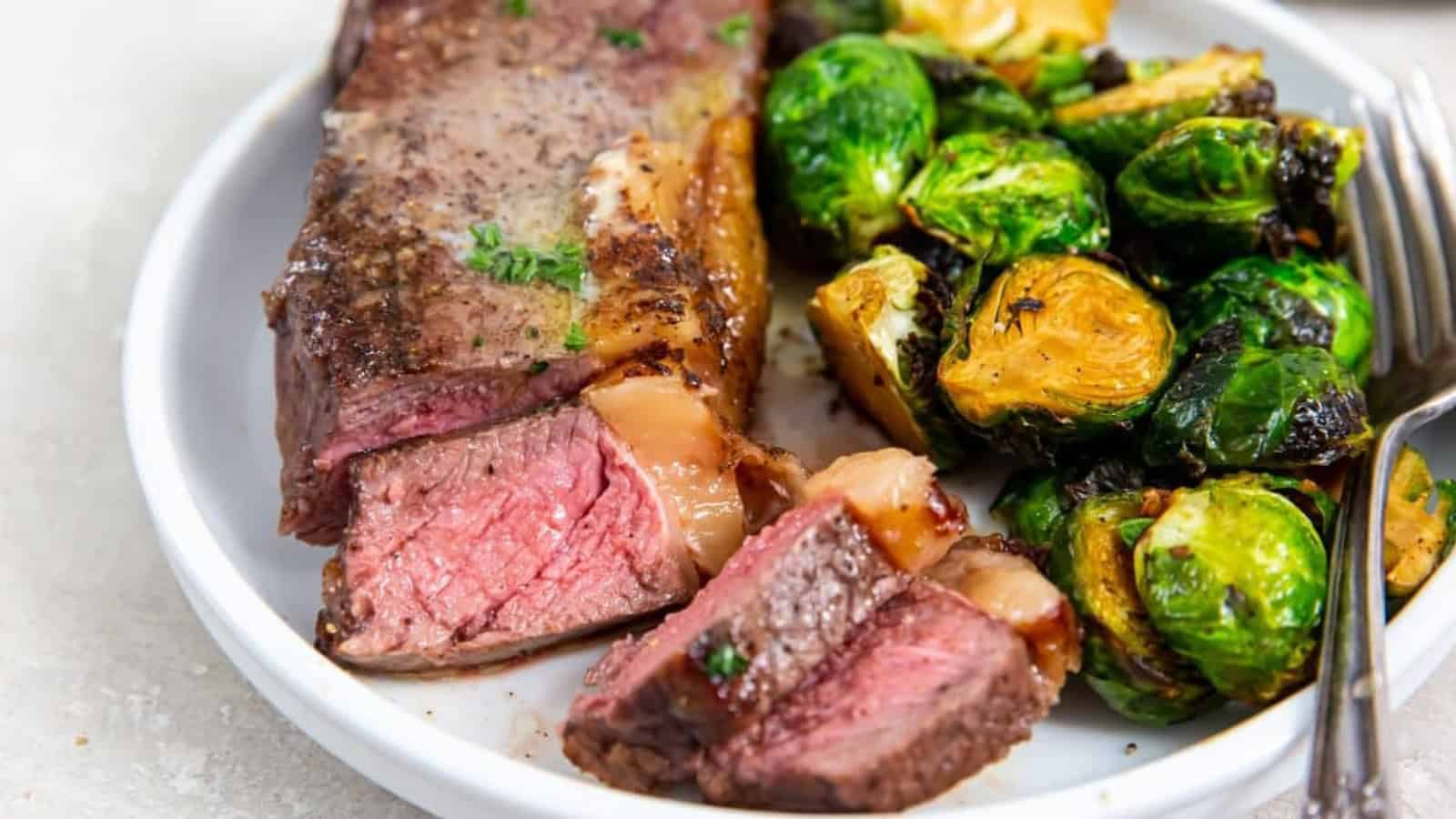 Air Fryer New York Strip Steaks on a white plate with butter, thyme, pepper, salt, brussels sprouts, and a fork.