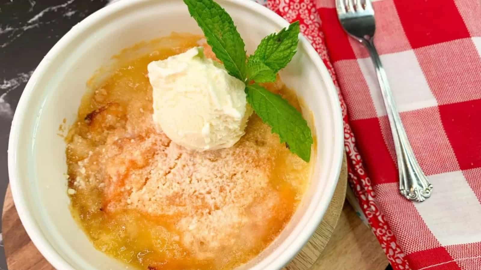 A bowl of peach cobbler topped with vanilla ice cream and a mint sprig, next to a fork and red napkin.