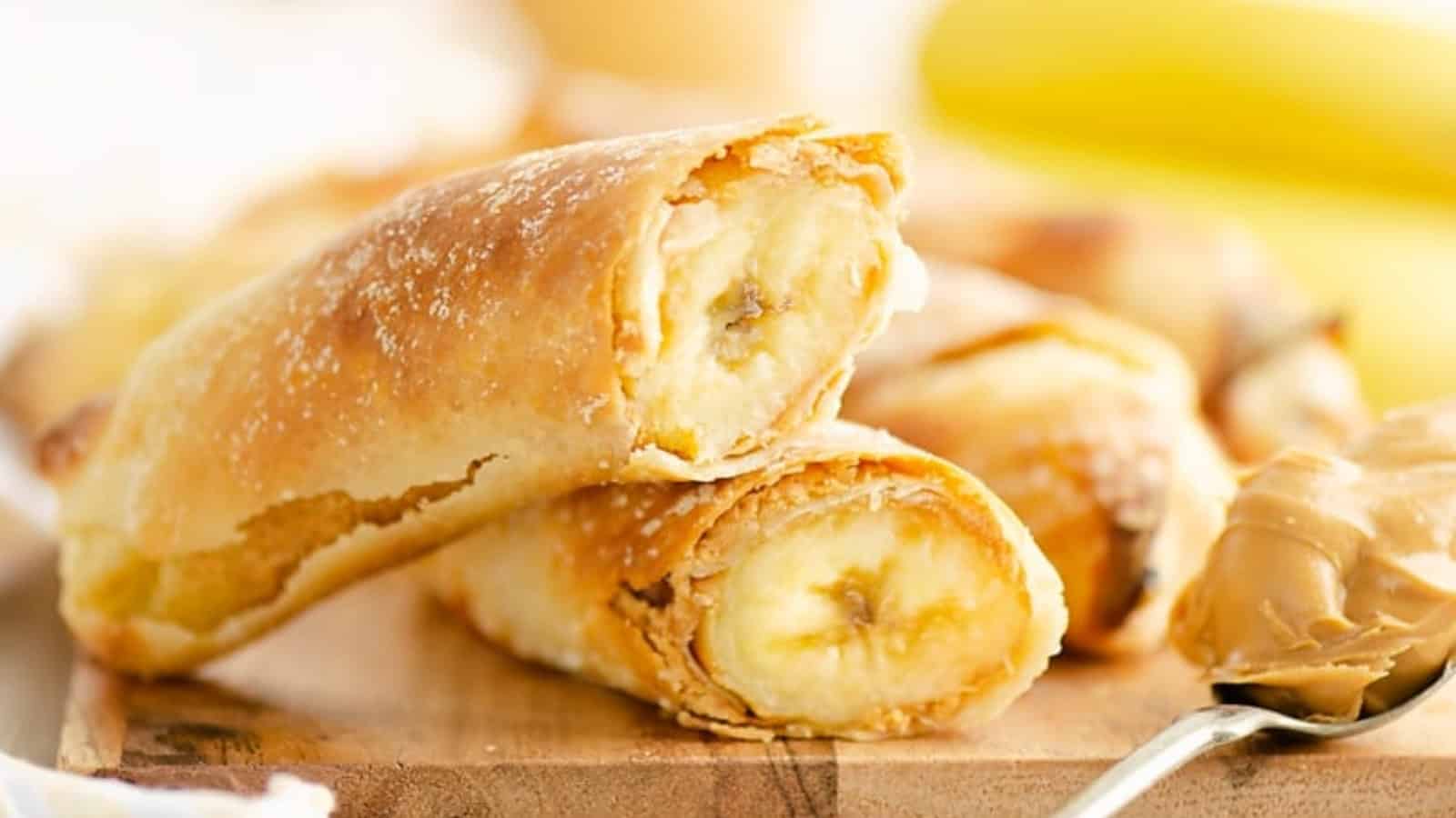 Close-up of banana-filled pastries on a wooden board, with a spoonful of peanut butter beside them.