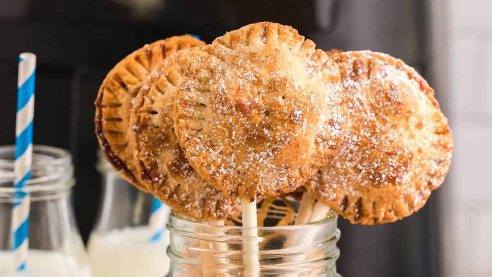 Round mini pie pops dusted with powdered sugar in a jar, with bottles of milk and striped straws in the background.