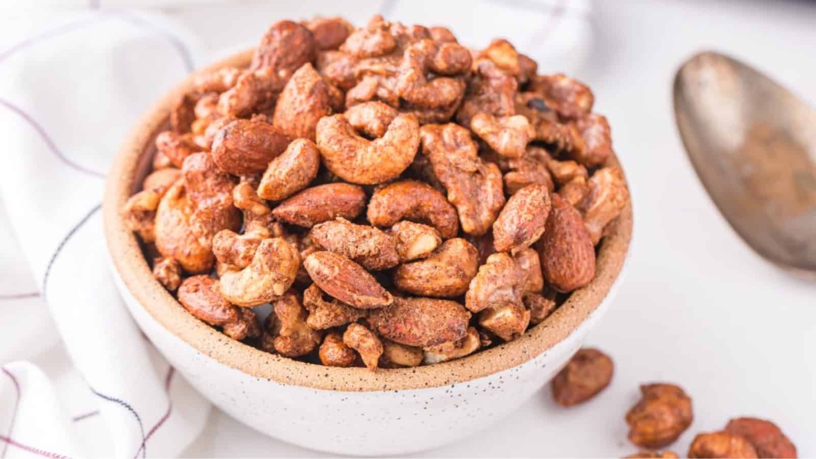 A white bowl filled with mixed roasted nuts sits on a white surface next to a spoon and a cloth.