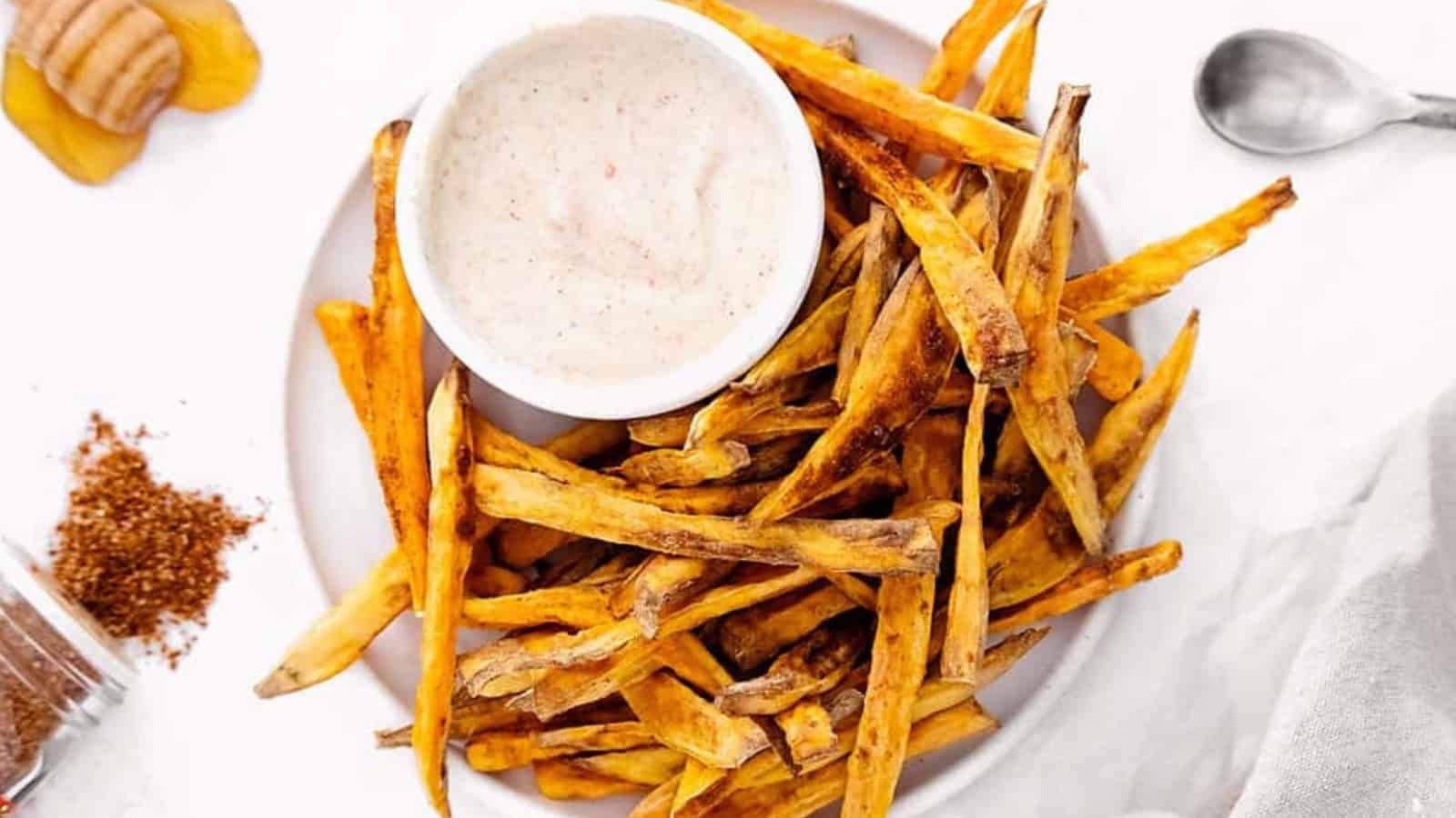 A plate of sweet potato fries with a bowl of dipping sauce on a white surface.