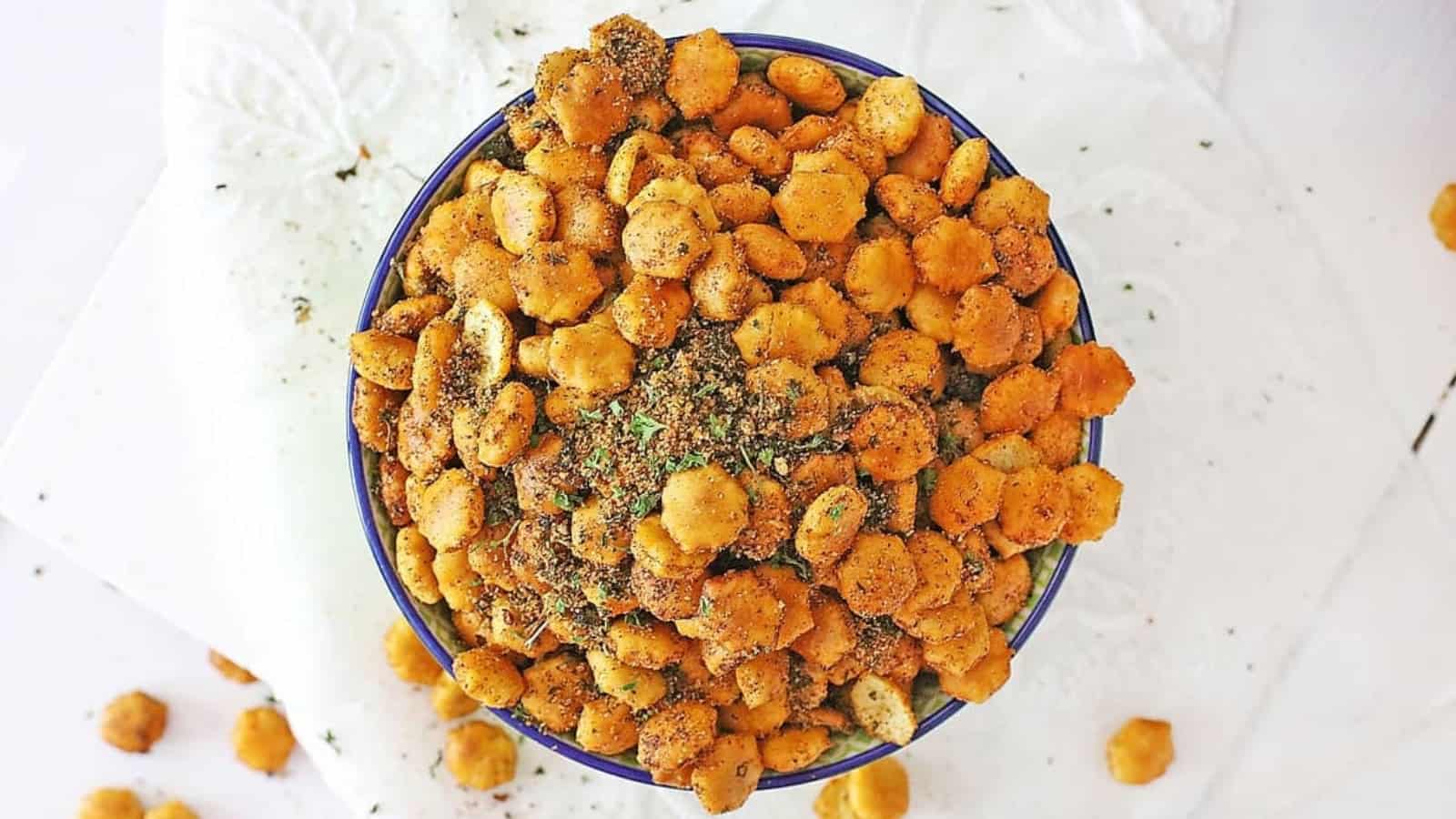 A bowl filled with seasoned, crunchy oyster crackers on a white surface with some crackers scattered around.