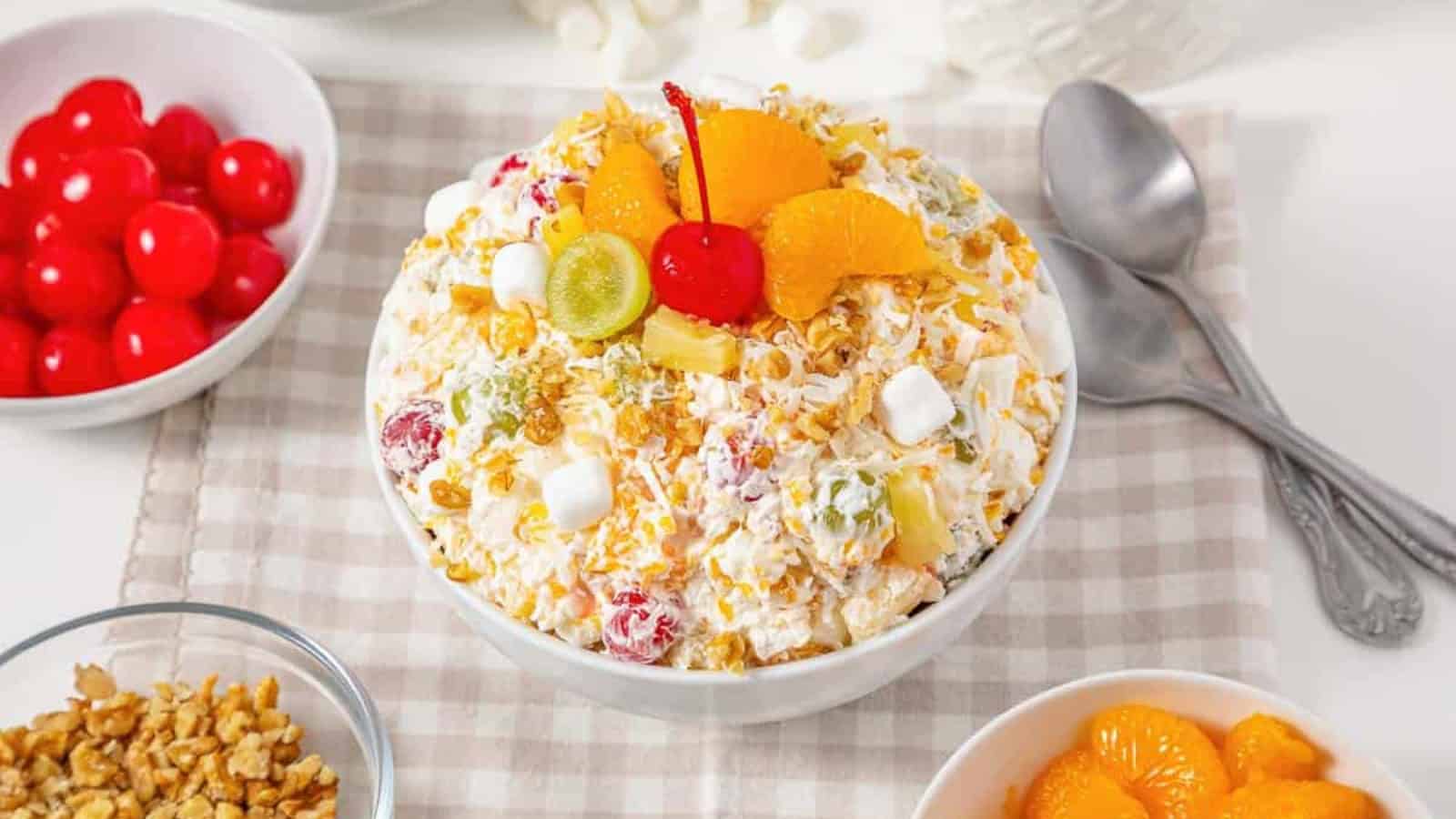 A bowl of ambrosia salad topped with fruit, marshmallows, and a cherry, with spoons and bowls of toppings nearby.