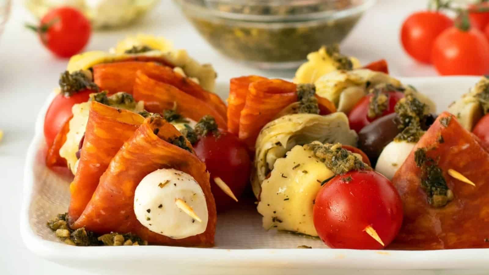 Antipasto skewers with pepperoni, mozzarella, tomatoes, olives, artichoke, and pesto on a white plate.