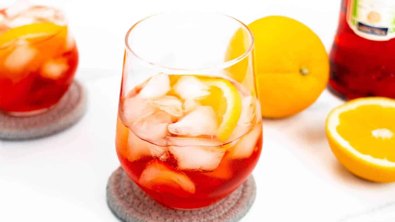 A glass of iced red cocktail with an orange slice, next to a whole and halved orange on a white surface.