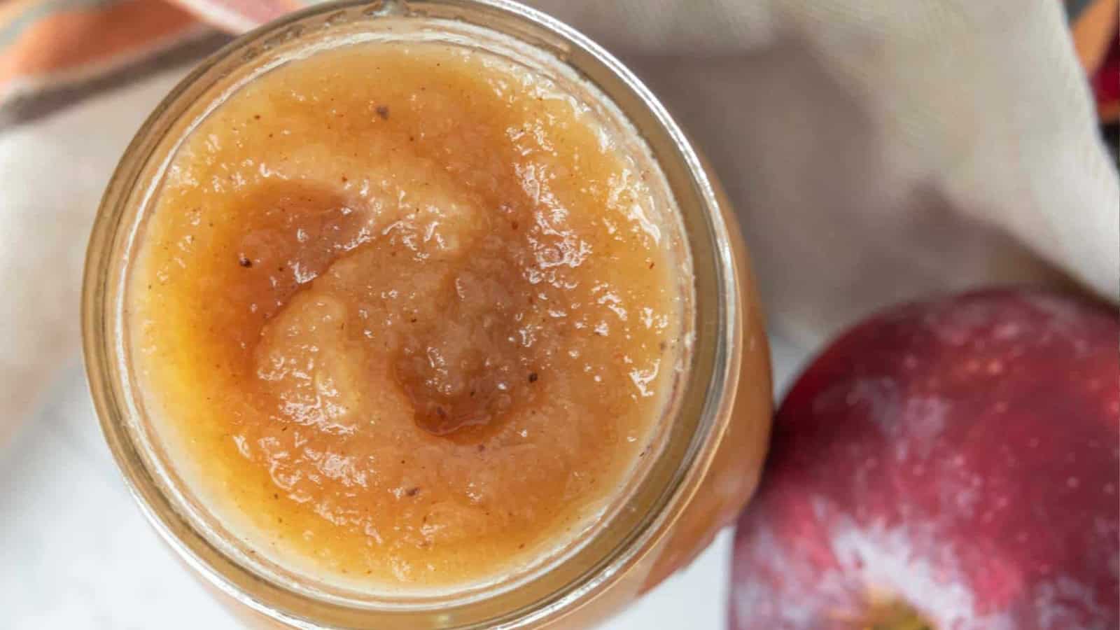 A jar of applesauce with visible spices next to a red apple on a light surface.