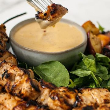 Grilled chicken skewers with greens and a bowl of dipping sauce, one piece being dipped with a fork.