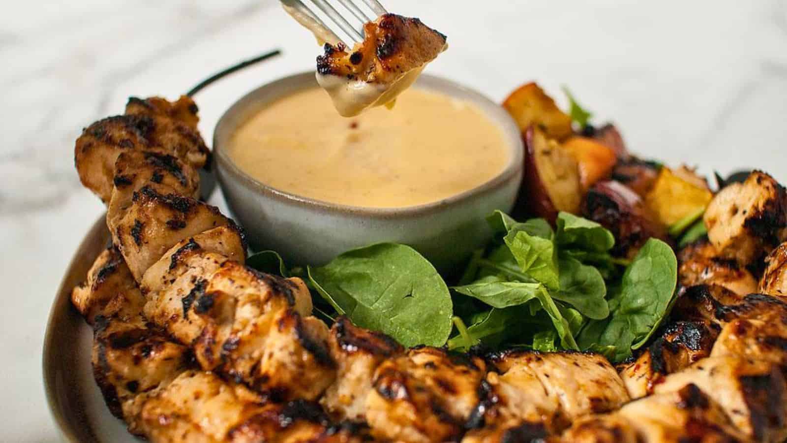 Grilled chicken skewers with greens and roasted veggies, served with a bowl of creamy dipping sauce.