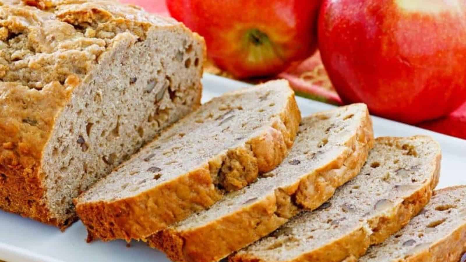 Sliced apple bread with visible nuts on a white plate, with whole red apples in the background.