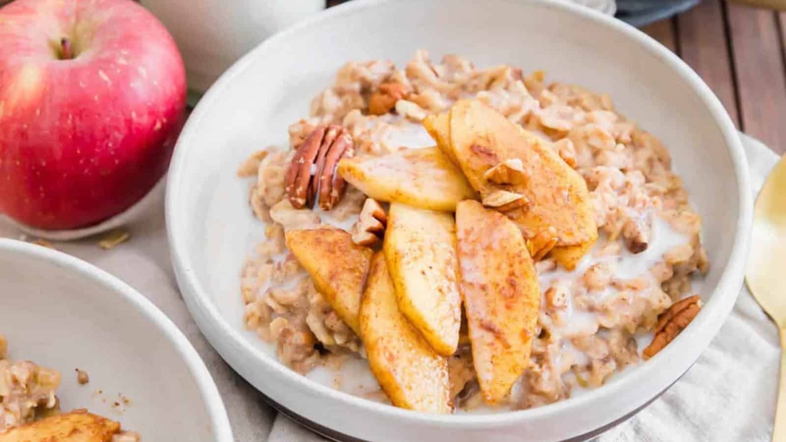 Bowl of oatmeal topped with sliced apples, pecans, and a splash of milk, with a red apple beside it.
