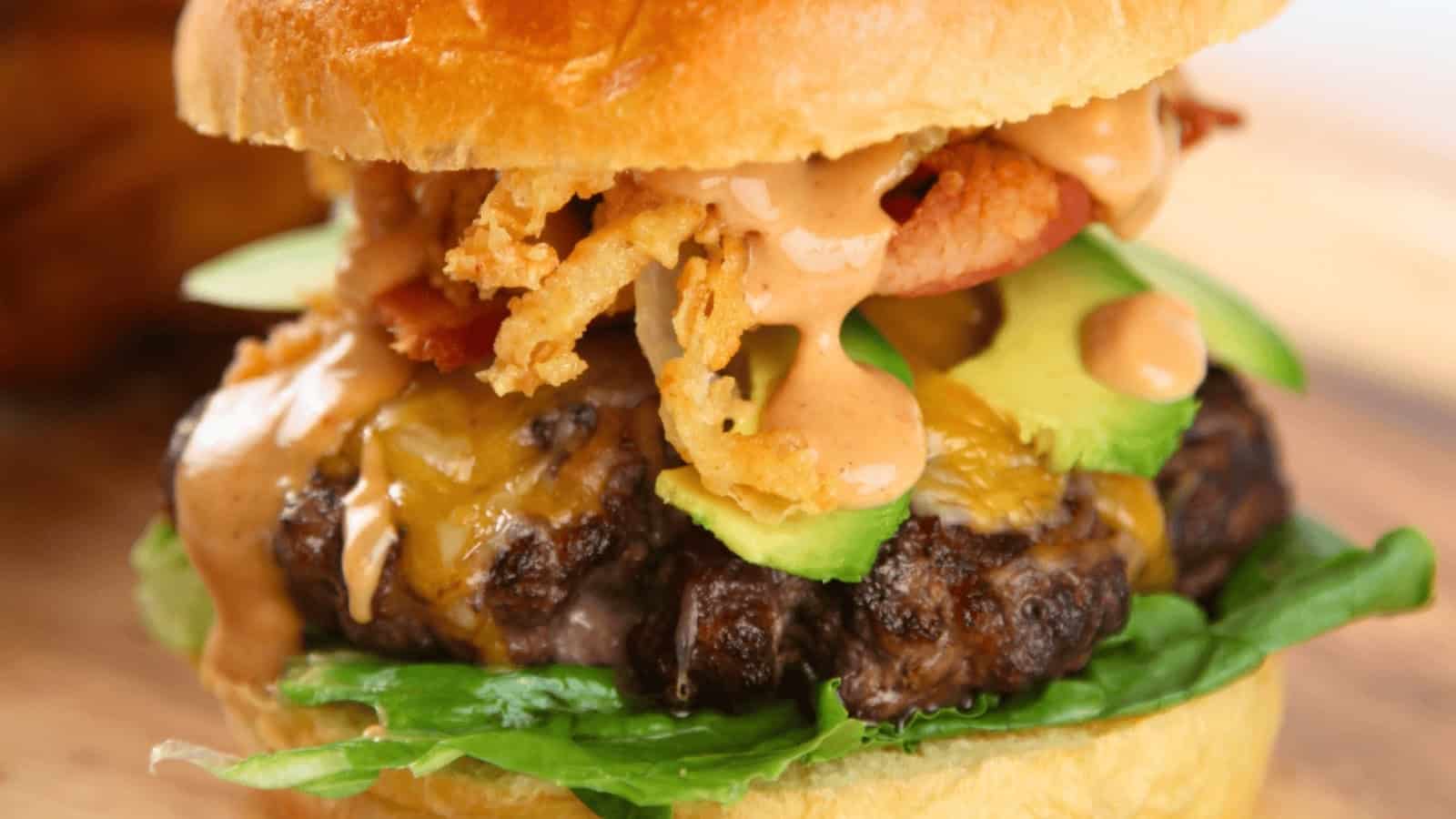 Close-up of a cheeseburger with lettuce, avocado, bacon, fried onions, and sauce on a toasted bun.